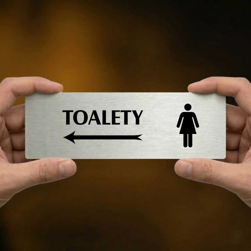 Toalety vlevo ženy, 240x79mm, stříbrná popiska s okrasným rámečkem